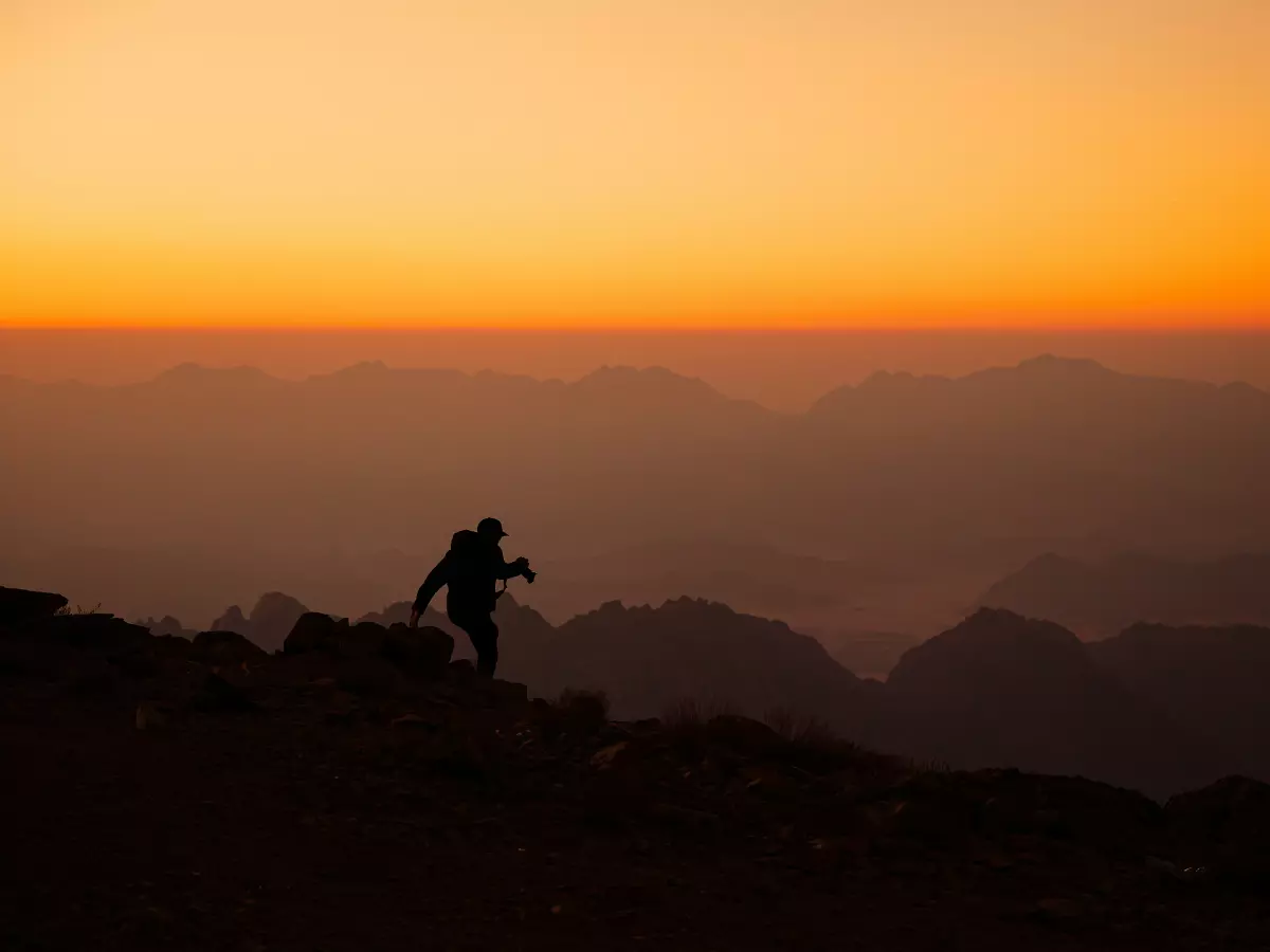 A silhouette of a person walking uphill in a mountain range, with the sunset in the background.