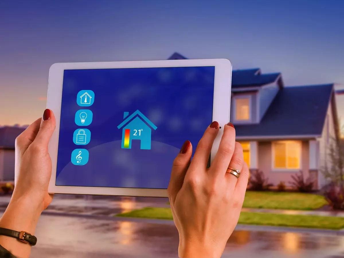 A woman's hands holding a tablet displaying a smart home app with icons for light control, temperature, door lock and music. The background is a blurred image of a modern house at dusk.