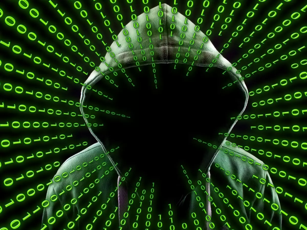 A hooded figure stands in front of a swirling background of binary code, their face obscured by the darkness. The green binary code represents the digital world of hacking and malware, while the hooded figure symbolizes the anonymity of the hacker.