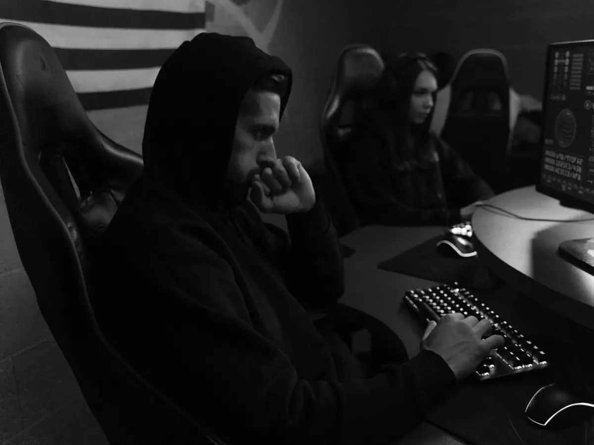 A person in a hoodie sits in a dimly lit room, hunched over a computer keyboard with a serious expression on their face, suggesting a focused and intense effort in the face of a cybersecurity challenge.