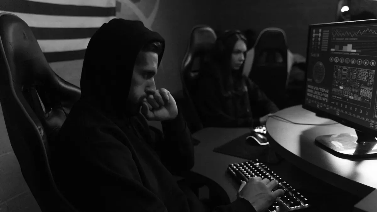 A person in a hoodie sits in a dimly lit room, hunched over a computer keyboard with a serious expression on their face, suggesting a focused and intense effort in the face of a cybersecurity challenge.