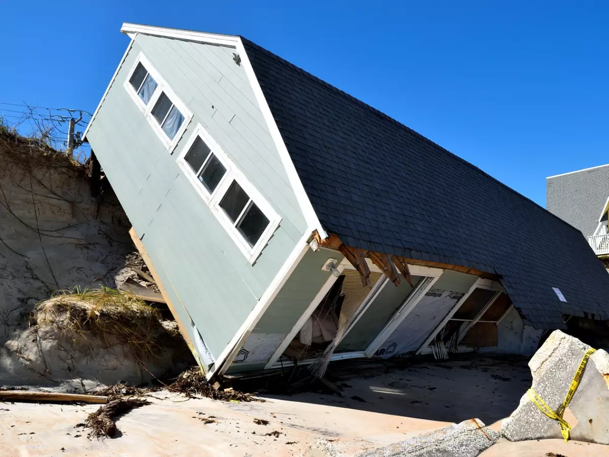 A house has been damaged by a hurricane. The house is tilted on its side and has a large hole in the roof. The house is surrounded by debris and sand.