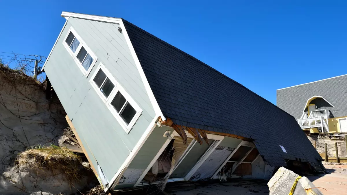 A house has been damaged by a hurricane. The house is tilted on its side and has a large hole in the roof. The house is surrounded by debris and sand.