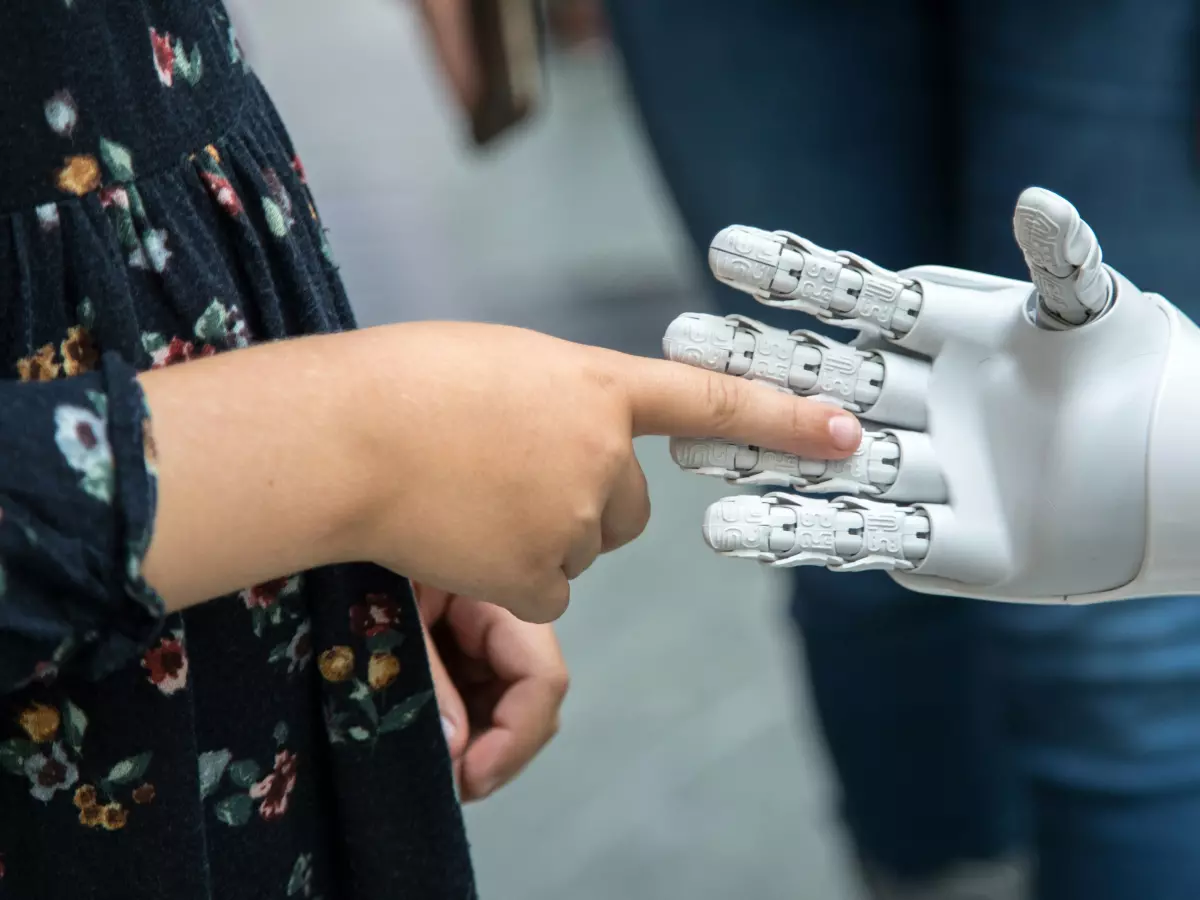 A close-up shot of a child's hand gently touching the fingers of a white robotic hand. The robotic hand is sleek and modern, showcasing its delicate touch.