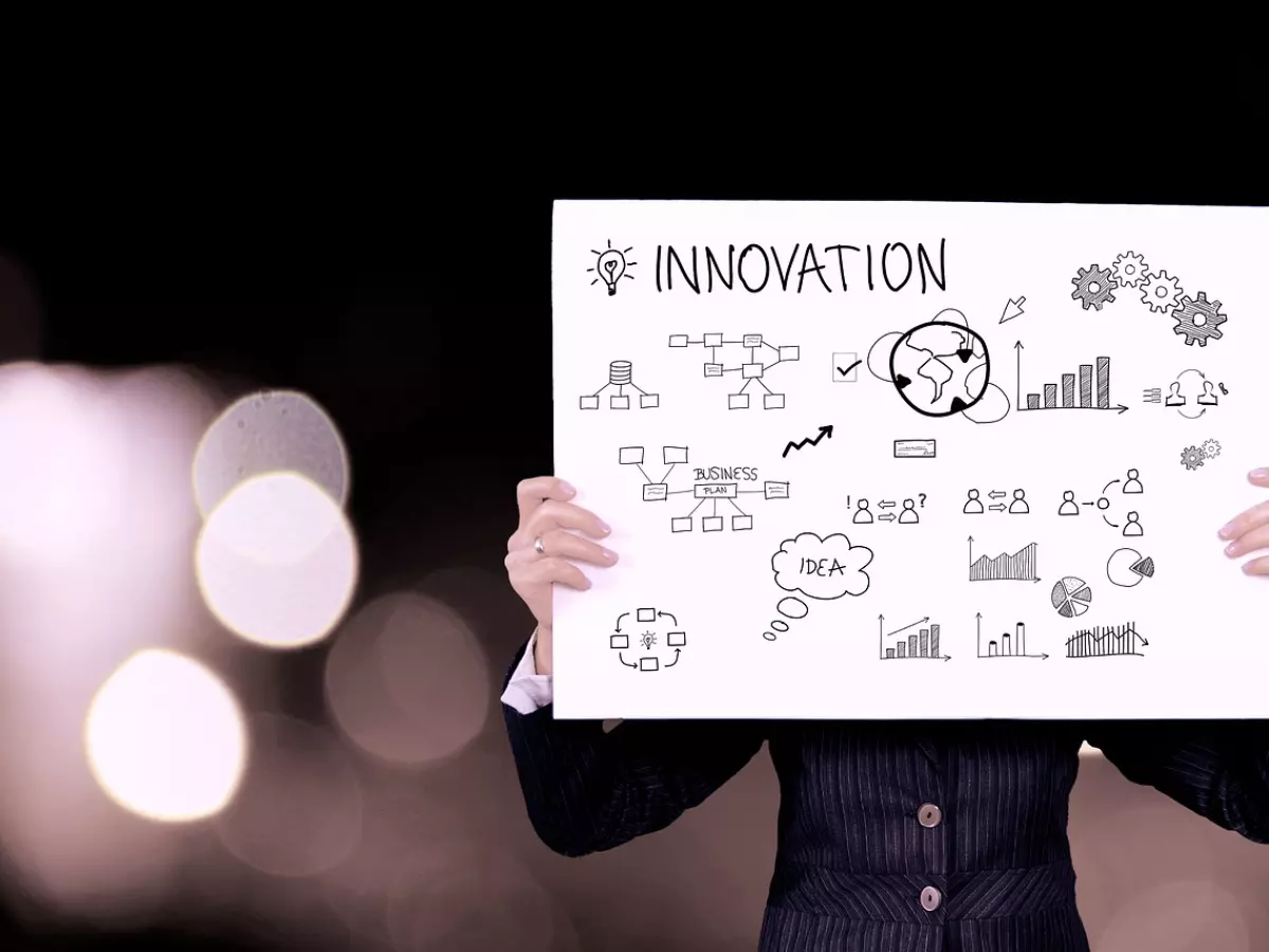 A person in a suit is holding a sign with the word "Innovation" written on it. The background is blurred and there are lights in the background. 