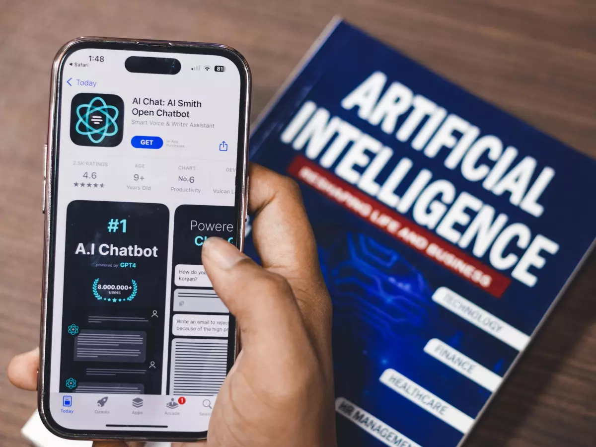 A hand holds a smartphone displaying an AI chatbot app on the screen, with a book titled 'Artificial Intelligence' visible in the background.