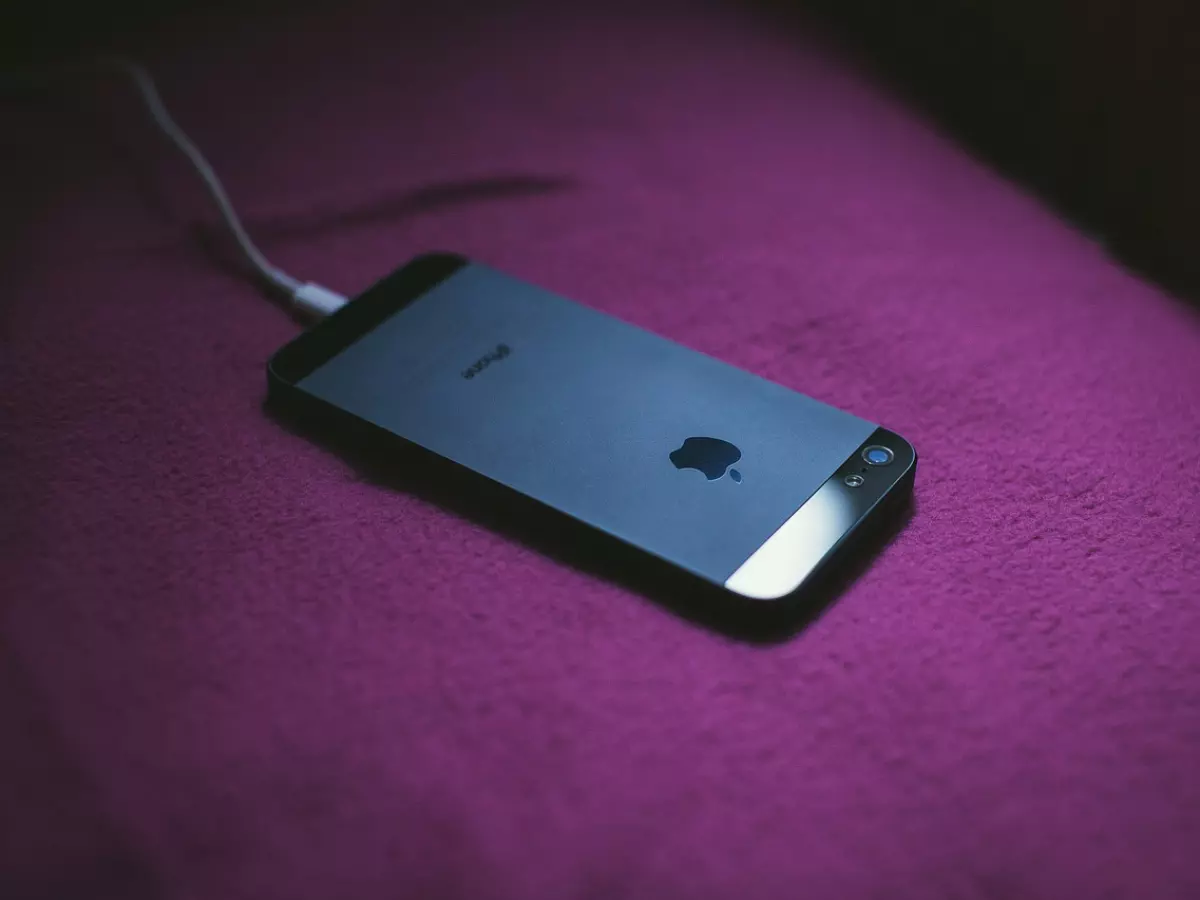 An Apple iPhone 5 is lying on a pink bed with a white charging cable connected.