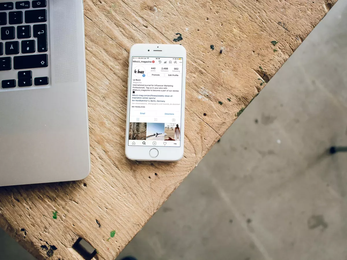 A white iPhone with a WhatsApp message on the screen sits on a wooden table next to a silver laptop. The background is a concrete floor.