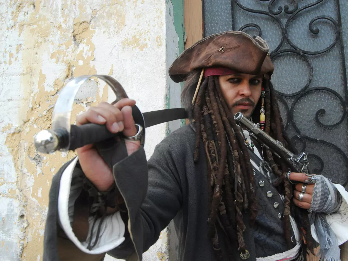 A man in a pirate costume, holding a sword and a gun, looking intense.