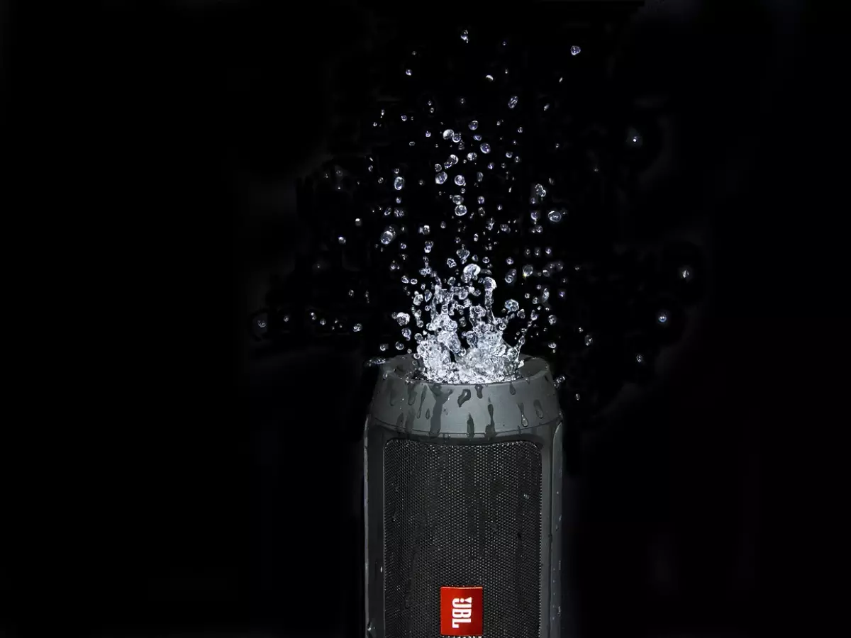 A black JBL speaker is submerged in water and water is spraying out of it. The speaker is on a black background.