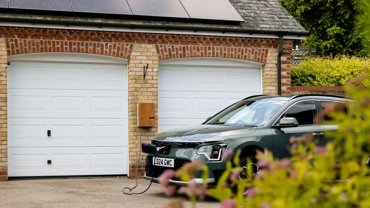 A dark green electric vehicle is plugged into a charging station in a garage with solar panels on the roof.