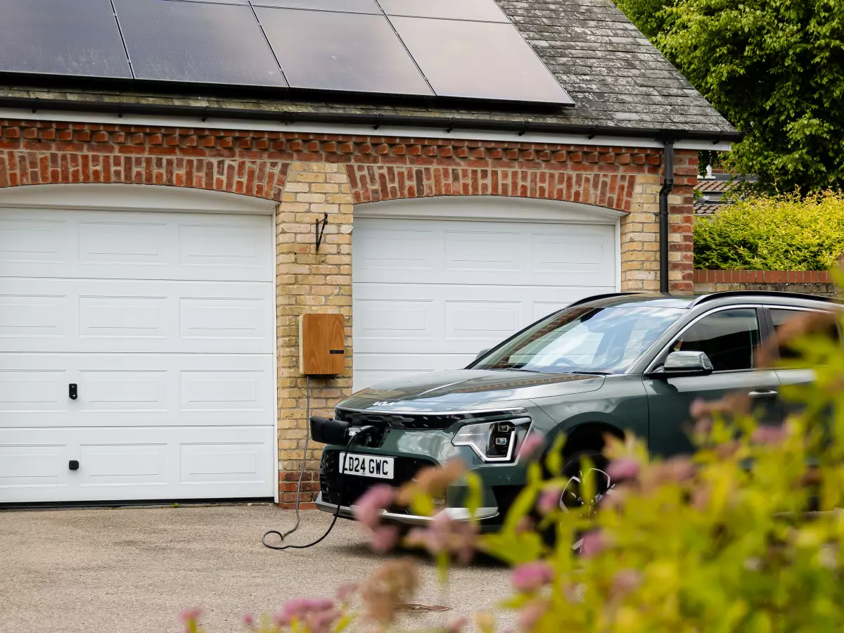 A dark green electric vehicle is plugged into a charging station in a garage with solar panels on the roof.