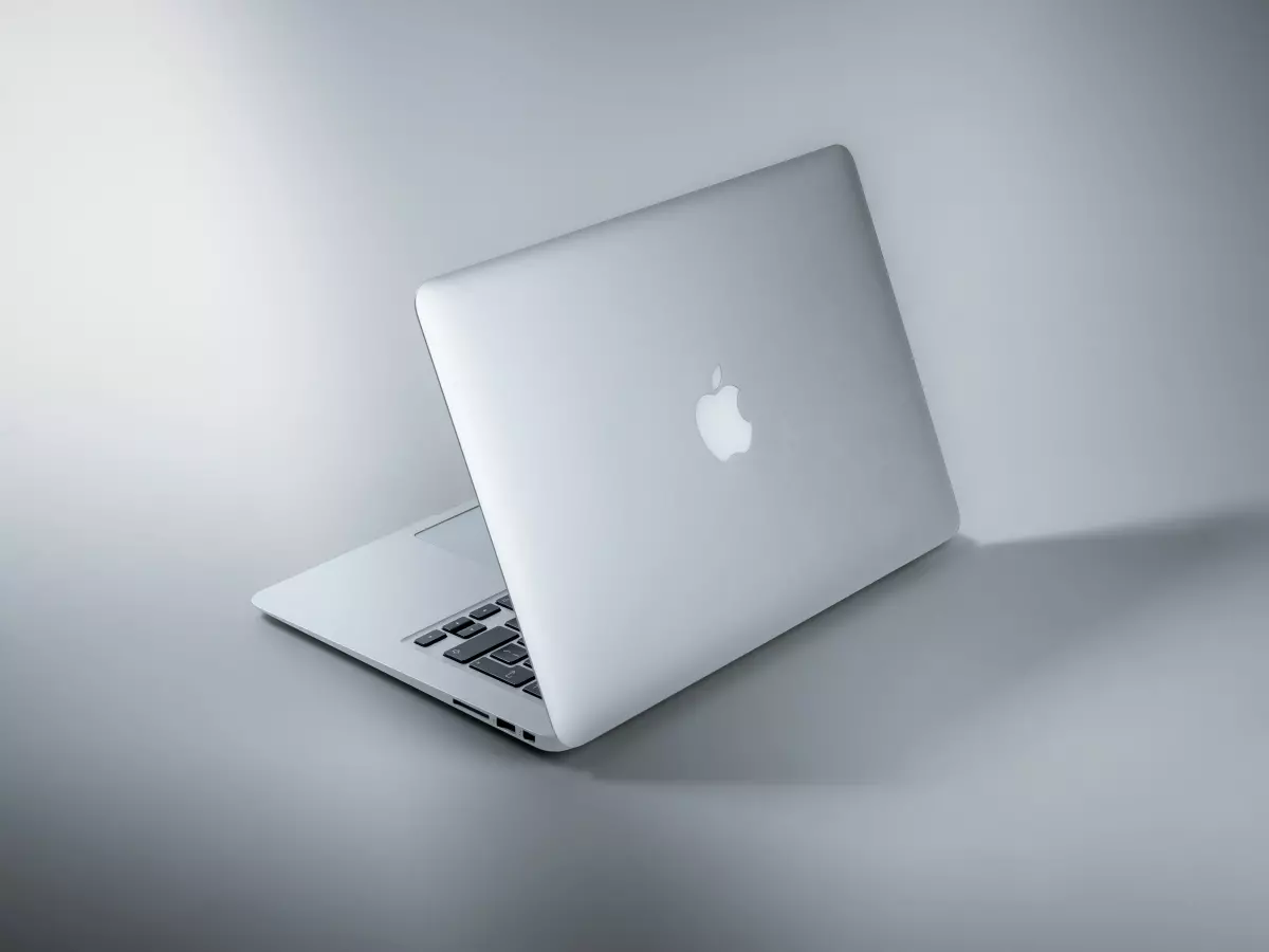 A white MacBook Air laptop is shown closed, with the lid slightly ajar. The laptop is resting on a white surface with a spotlight illuminating it from the left side. The Apple logo is visible on the closed lid.