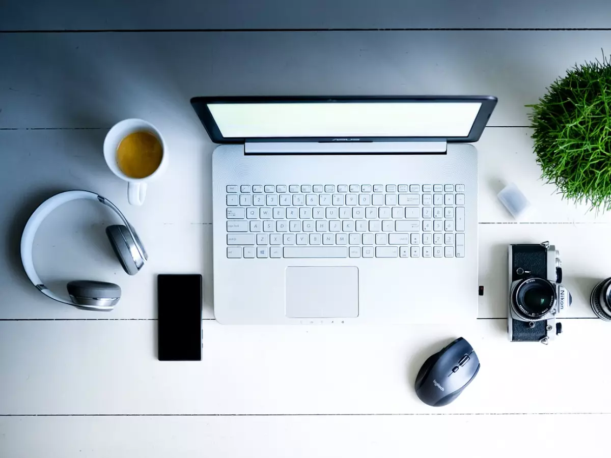 A white laptop, a smartphone, headphones, a camera, a mouse, a cup of coffee and a plant on a white wooden table.