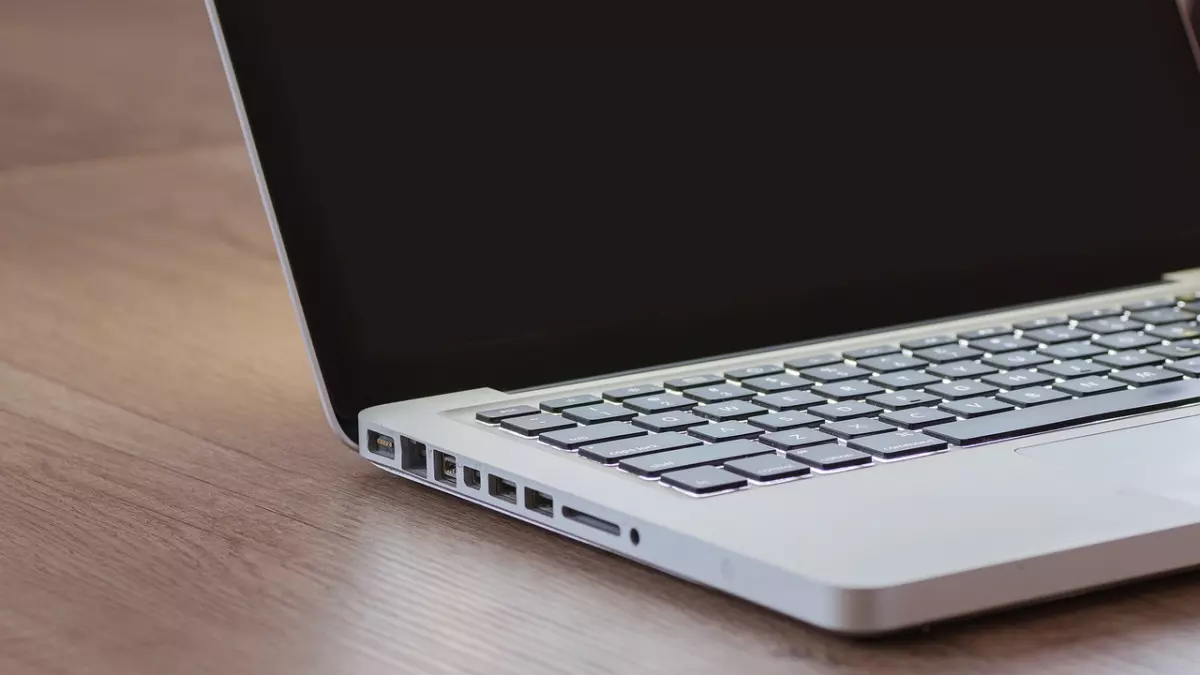 A silver MacBook laptop rests on a wooden table, seen from the side with the screen closed. The keyboard and ports are visible, highlighting its sleek design and functionality.