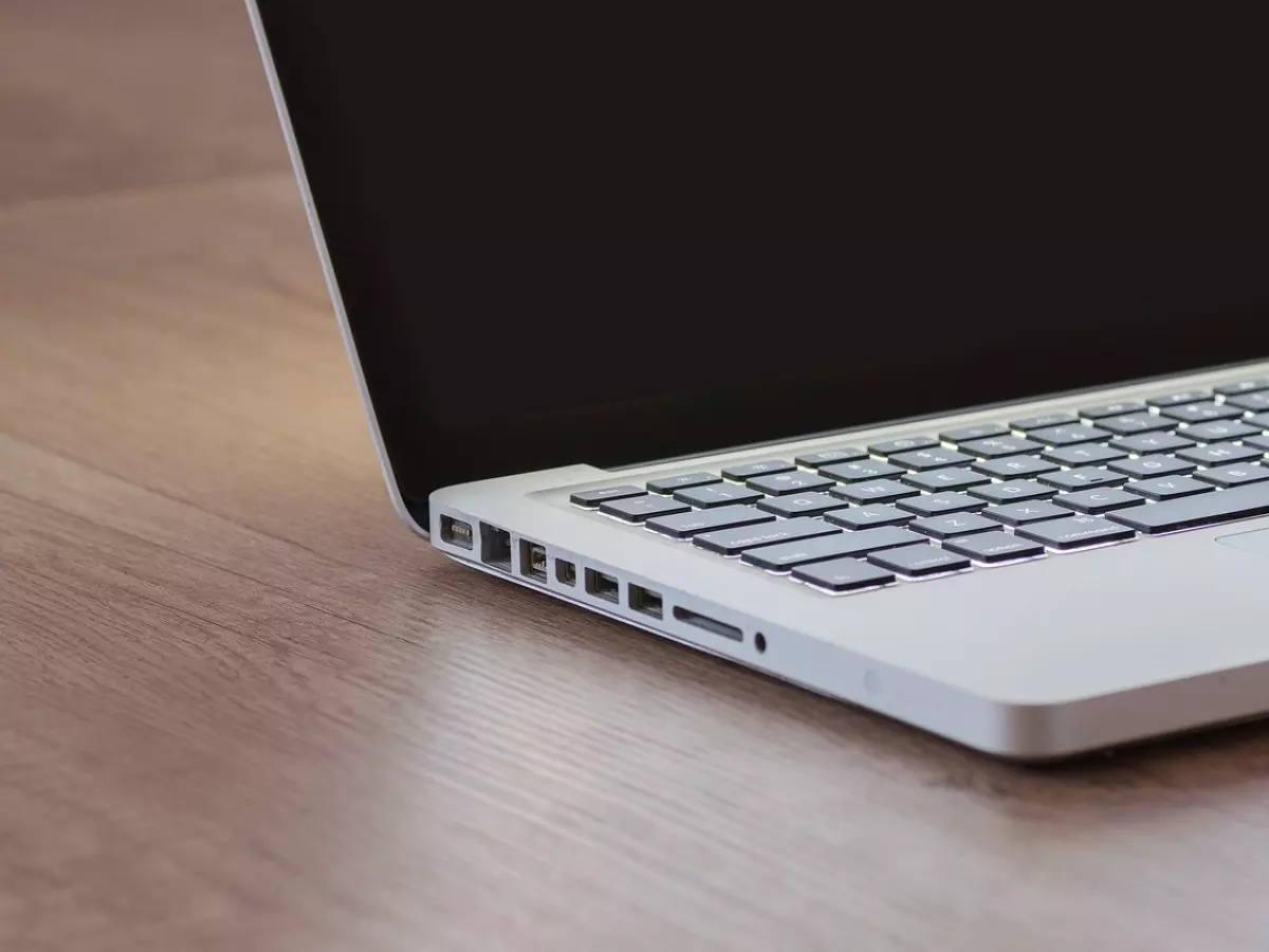 A silver MacBook laptop rests on a wooden table, seen from the side with the screen closed. The keyboard and ports are visible, highlighting its sleek design and functionality.