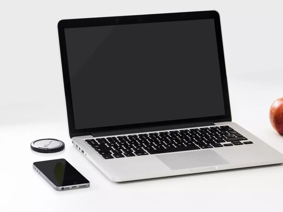 A silver laptop with a black screen, a smartphone, a camera lens cap, and a red apple on a white table.