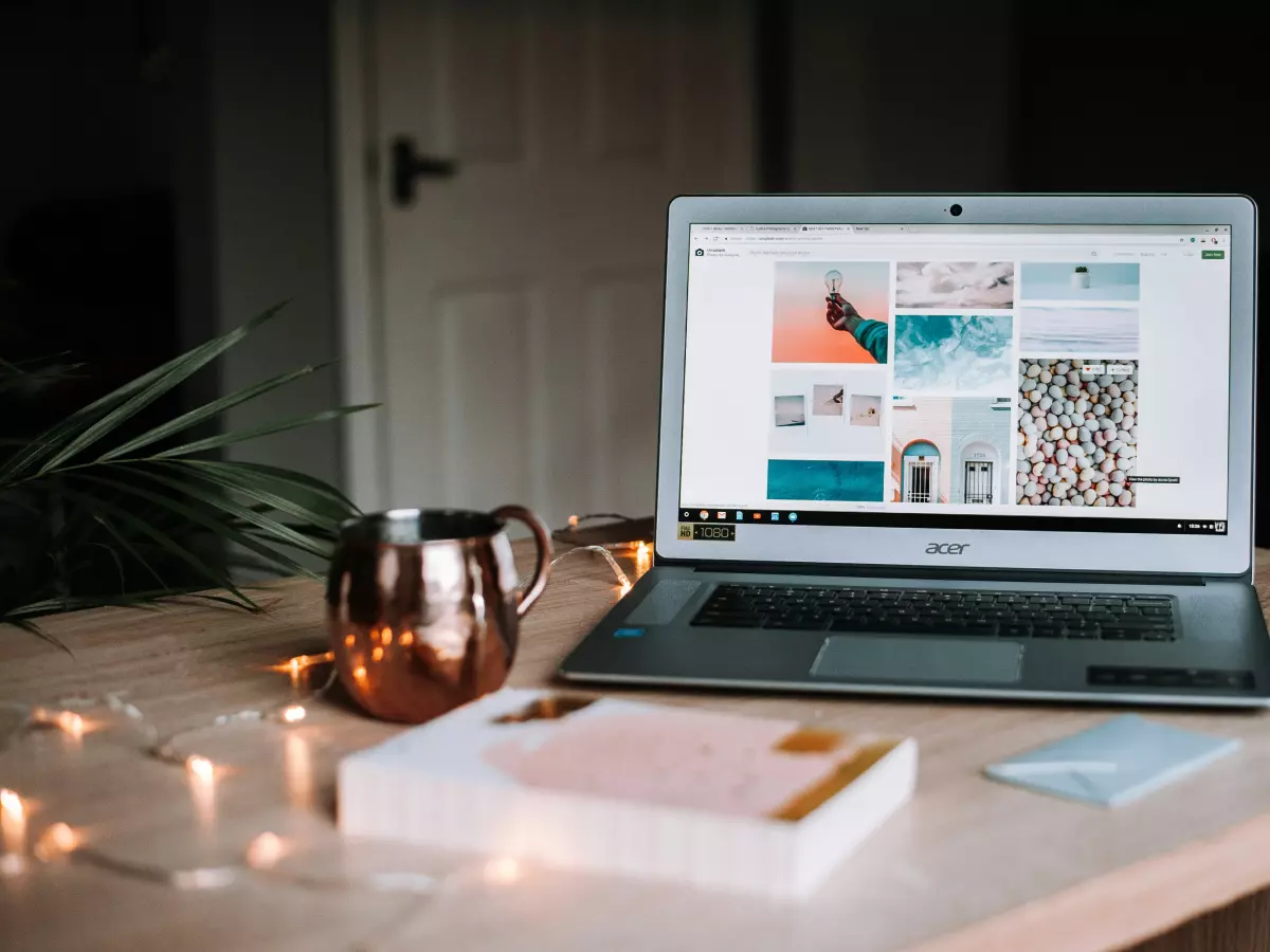 A laptop is open on a wooden table. It has a light background, showing a website. A copper mug and a book are also on the table, along with string lights.