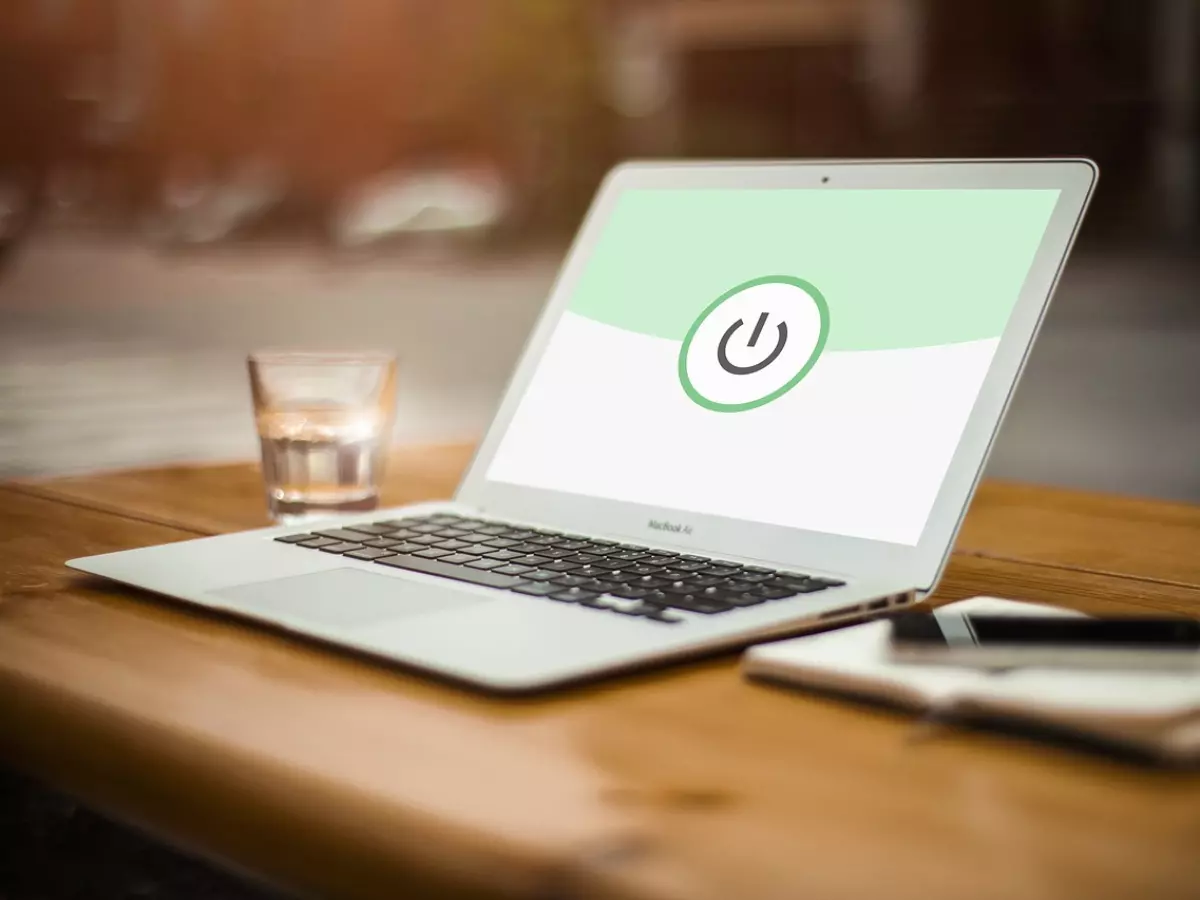 A laptop with a power button on the screen and a glass of water on a table with a notepad and a phone.