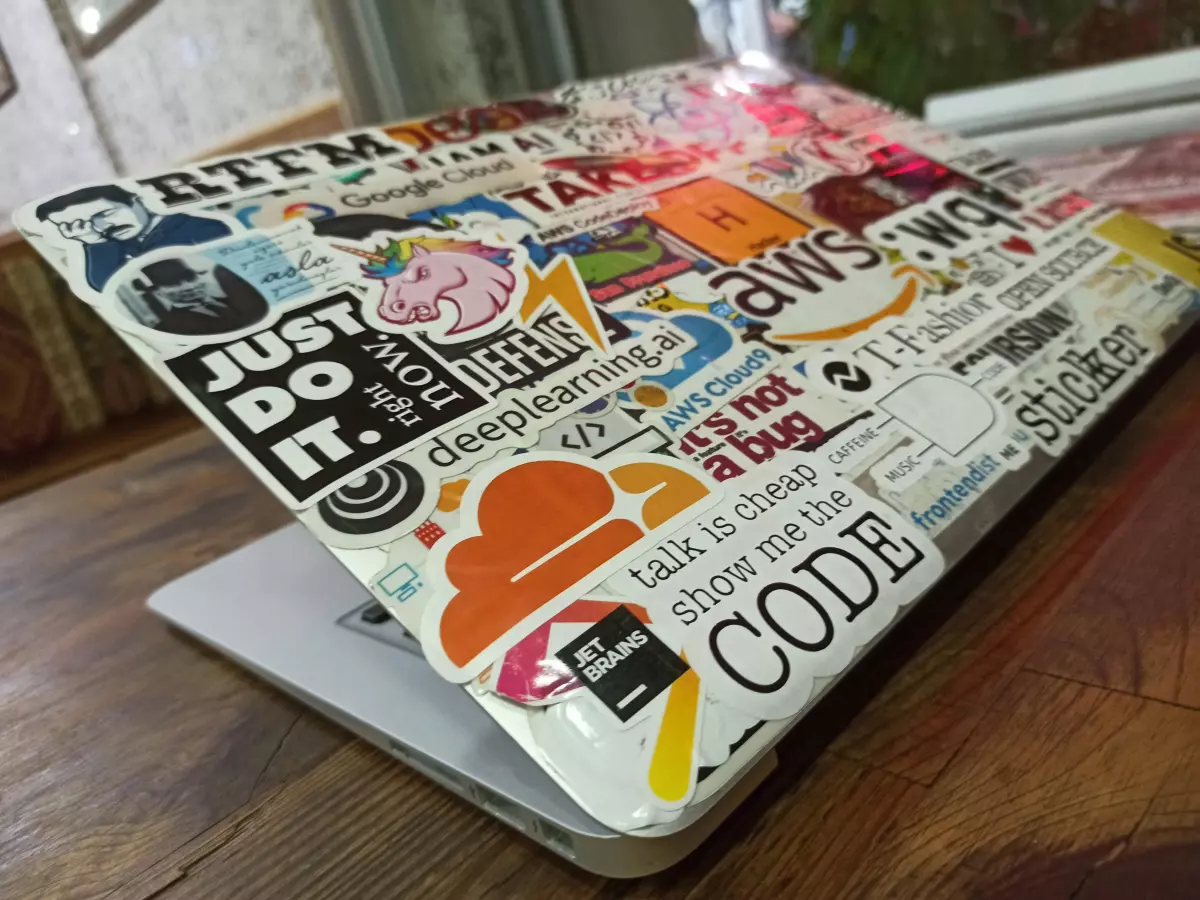 A laptop covered with stickers, including one with the AWS logo. The stickers feature tech-related themes and phrases.