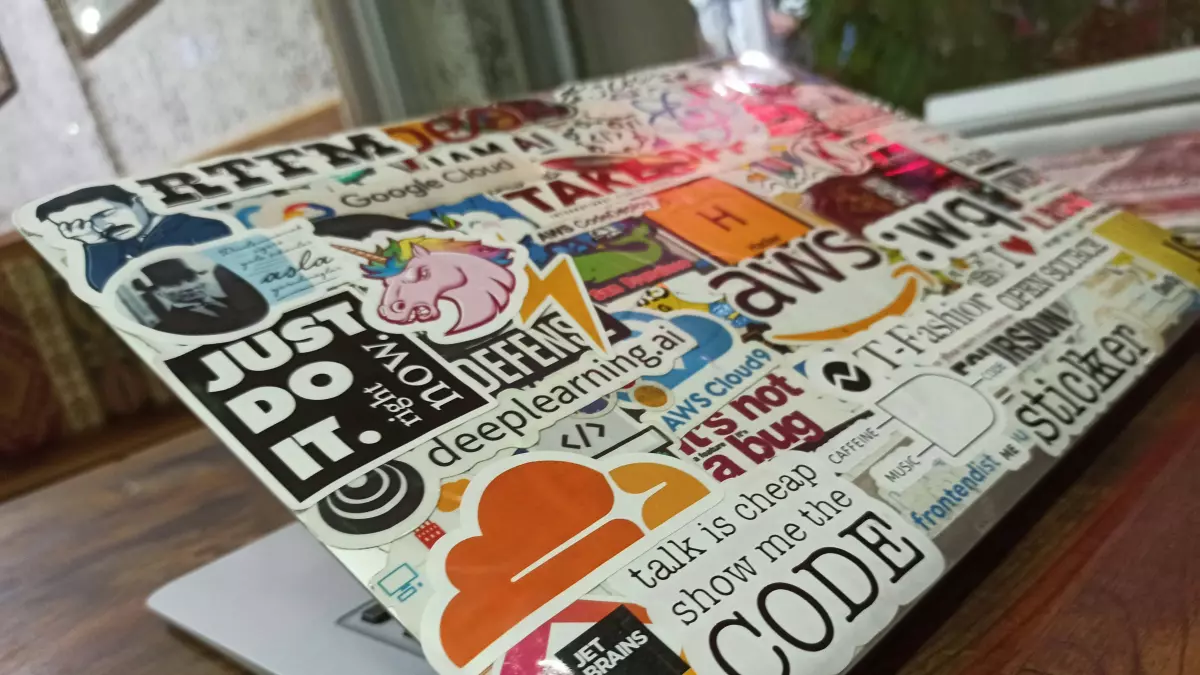 A laptop covered with stickers, including one with the AWS logo. The stickers feature tech-related themes and phrases.