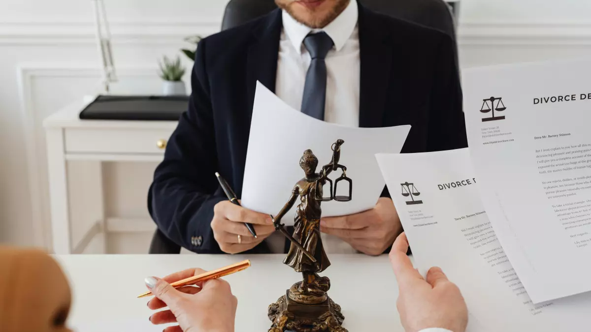 A lawyer in a suit is sitting at a desk reviewing legal documents with a client. A statue of justice sits between them. The image conveys a sense of legal consultation and serious discussions.