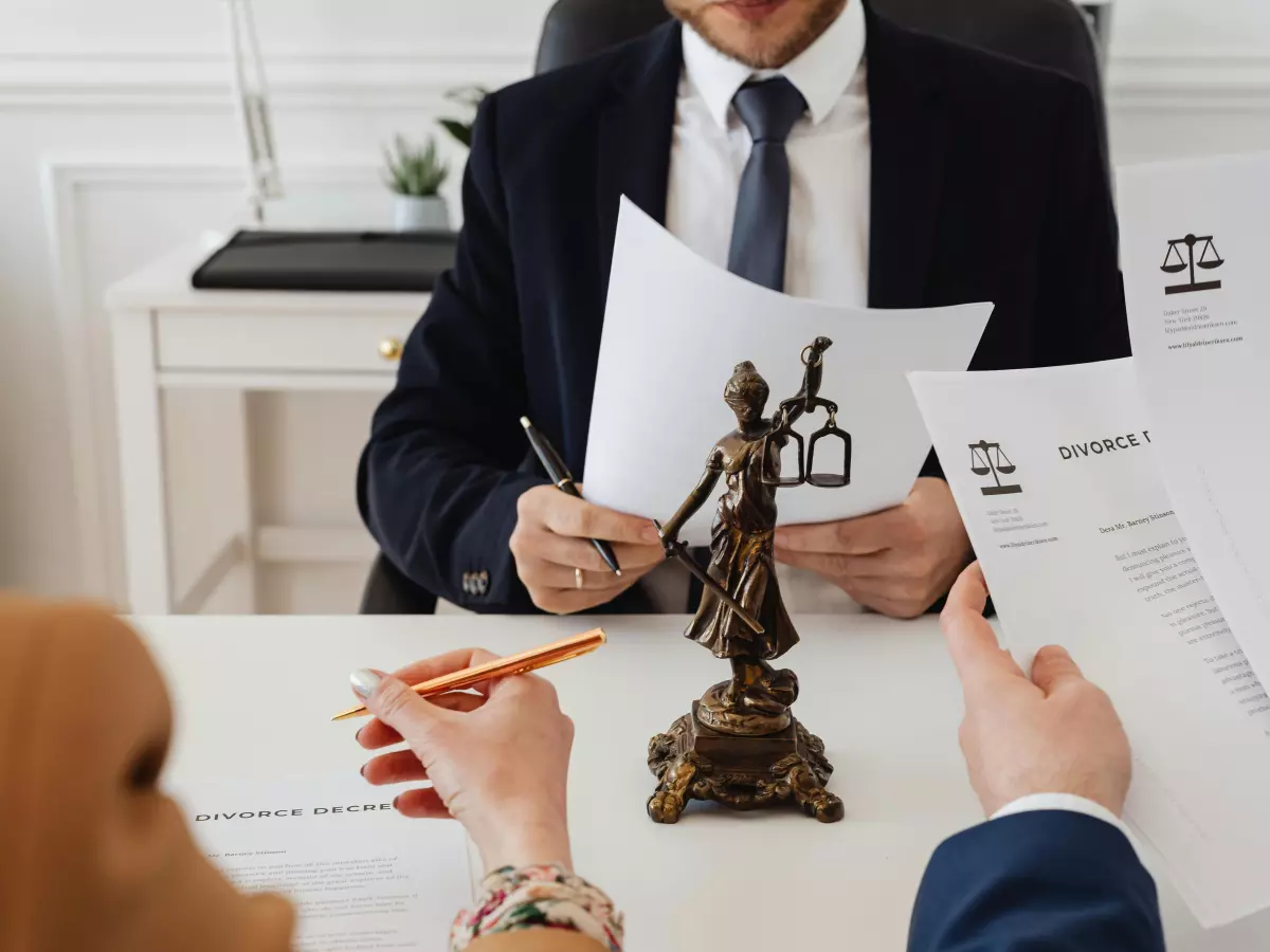 A lawyer in a suit is sitting at a desk reviewing legal documents with a client. A statue of justice sits between them. The image conveys a sense of legal consultation and serious discussions.