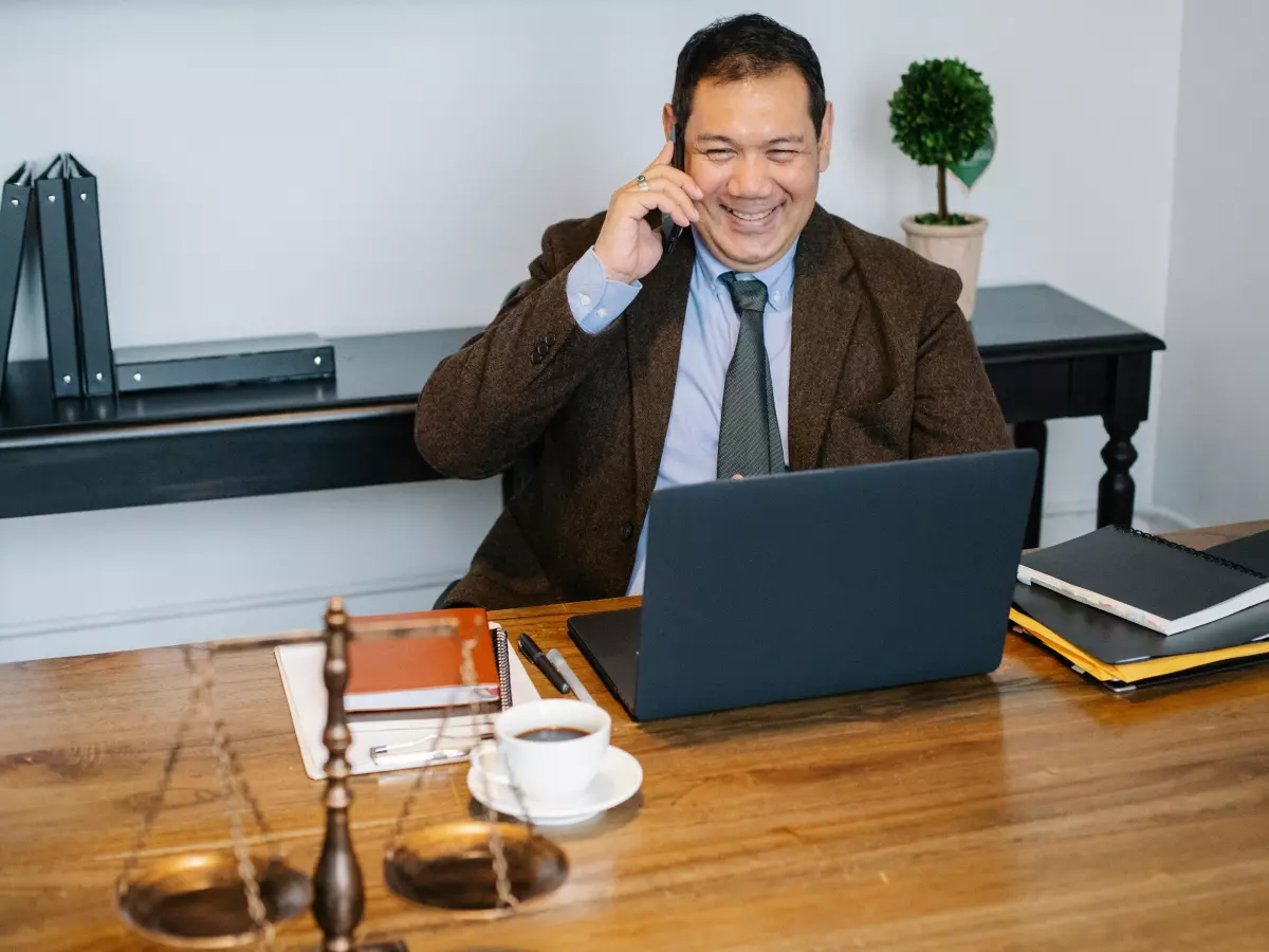 A man in a suit sitting at a desk with a laptop and a phone. He is talking on the phone and smiling.