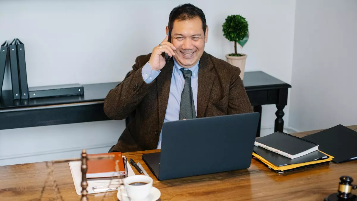 A man in a suit sitting at a desk with a laptop and a phone. He is talking on the phone and smiling.