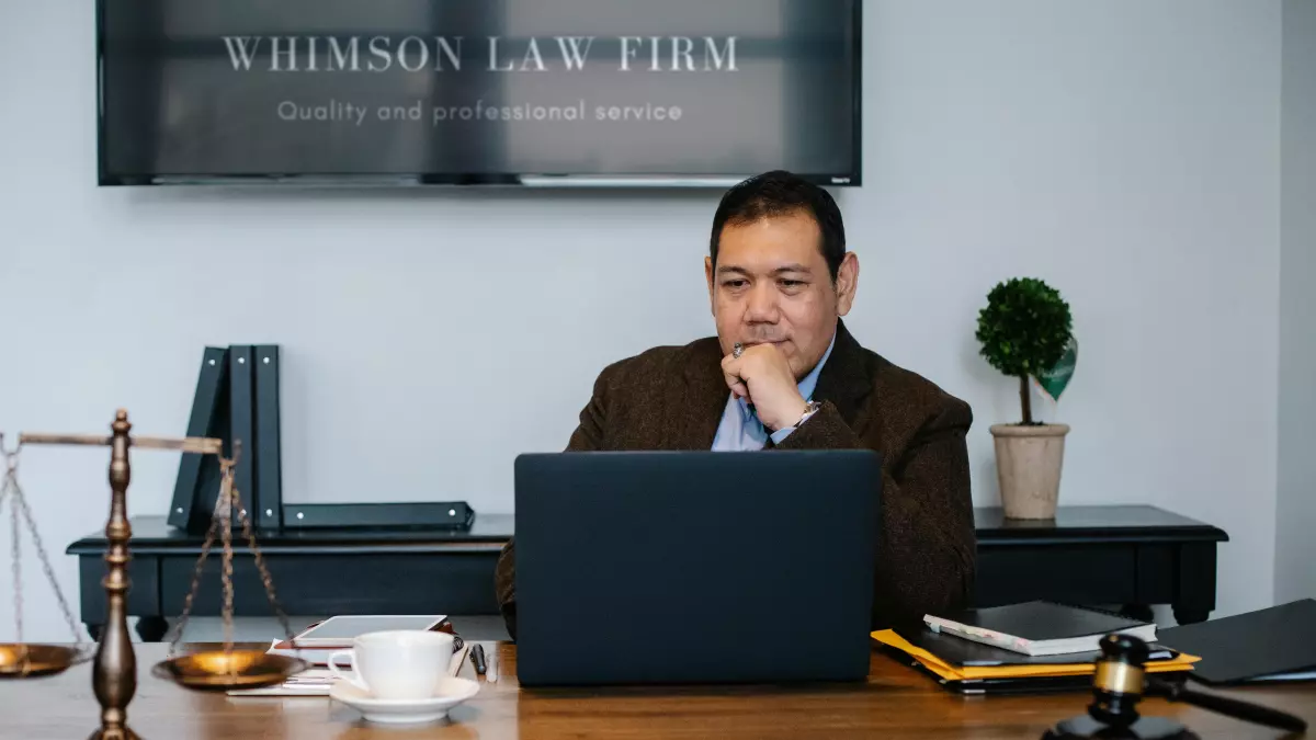 A man sitting at a desk in a suit, looking at a laptop computer with a scale and a gavel in the background.