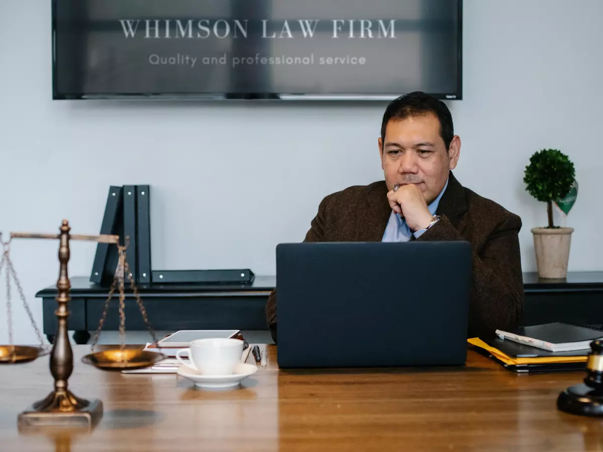 A man sitting at a desk in a suit, looking at a laptop computer with a scale and a gavel in the background.