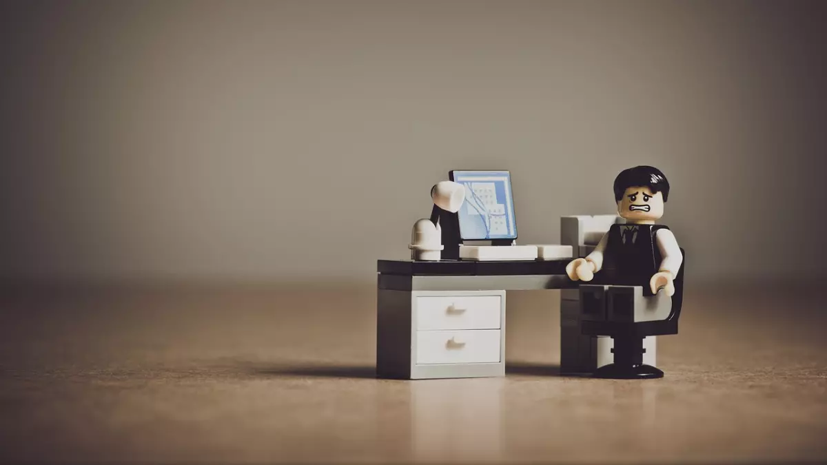 A miniature Lego figurine of a person, dressed as a businessman, is sitting in a chair at a Lego desk, looking distressed. He is seated in front of a Lego computer.