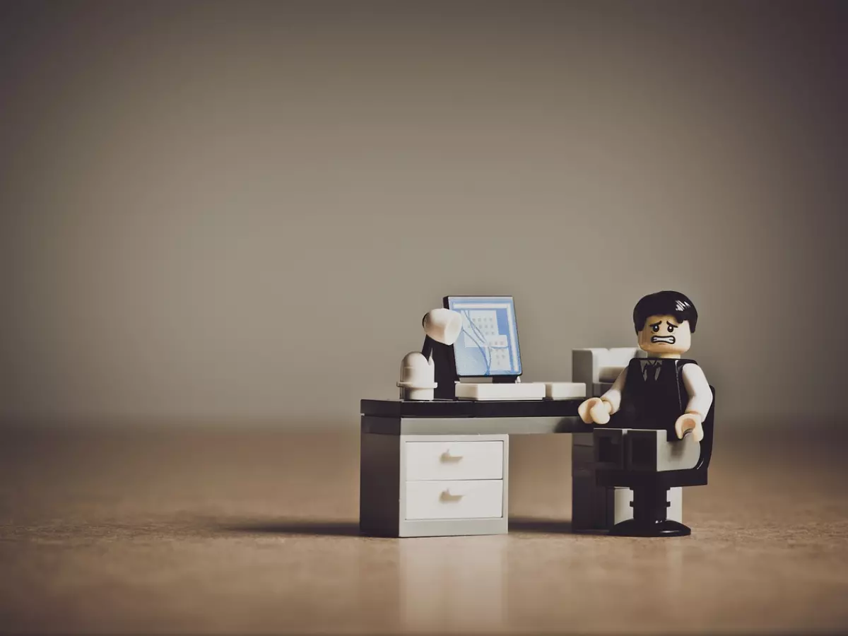 A miniature Lego figurine of a person, dressed as a businessman, is sitting in a chair at a Lego desk, looking distressed. He is seated in front of a Lego computer.