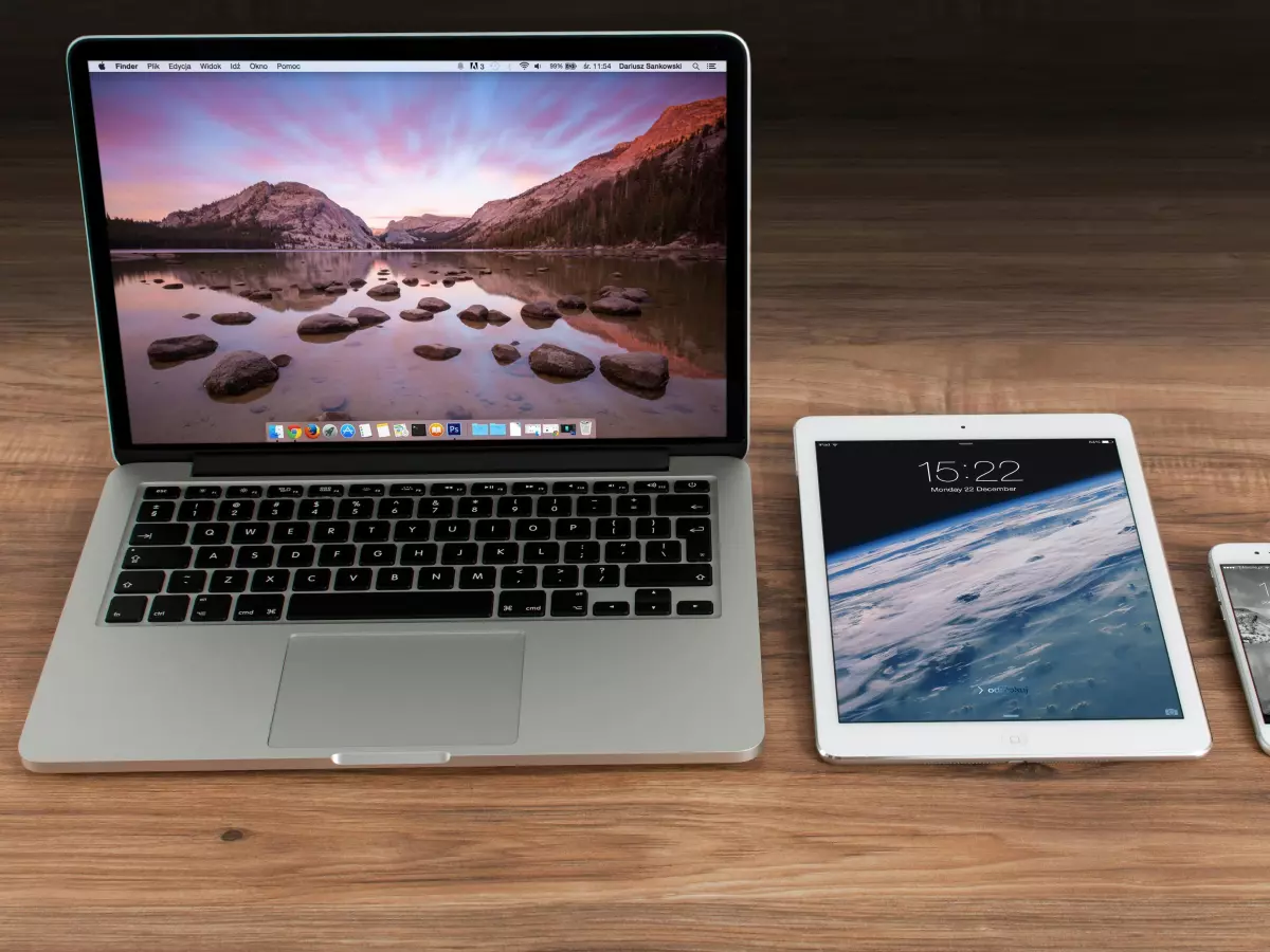 An iPhone, iPad, and MacBook are lined up on a wooden surface. The iPad is showing a beach scene, and the iPhone is showing a clock.