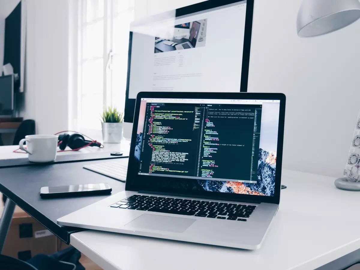 A laptop is open on a desk, with a cup of coffee and headphones next to it. The screen is displaying code. There is a second monitor behind the laptop, showing a website.