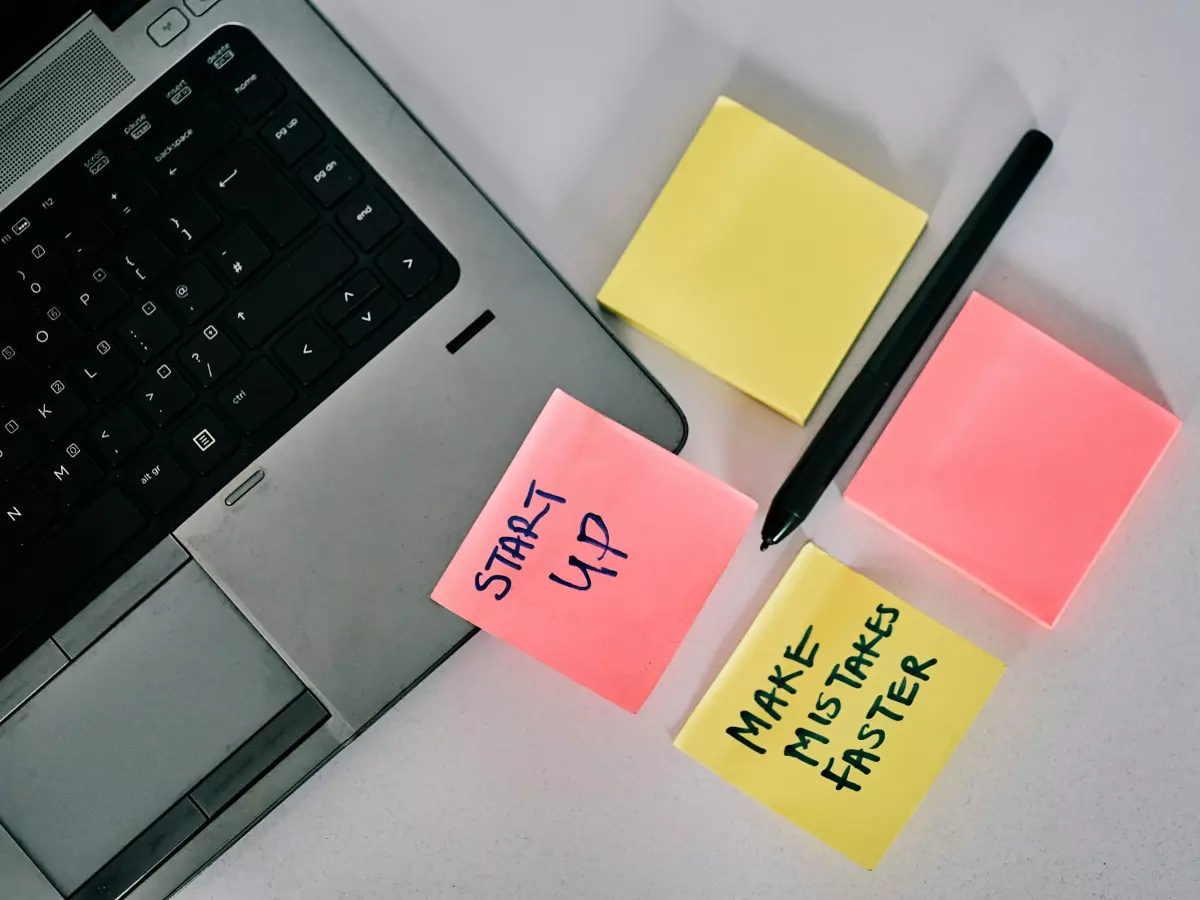 A laptop with sticky notes on a white surface, The sticky notes say "start up", "make mistakes faster" and one is blank.