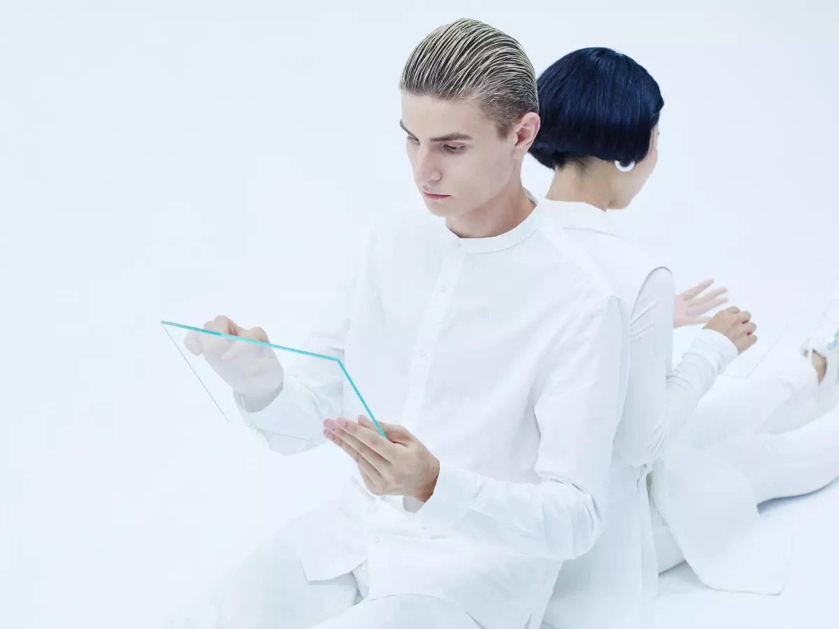 Two people in white clothing, one sitting and one lying down, in a white room. The sitting person is holding a small white device in their hands.