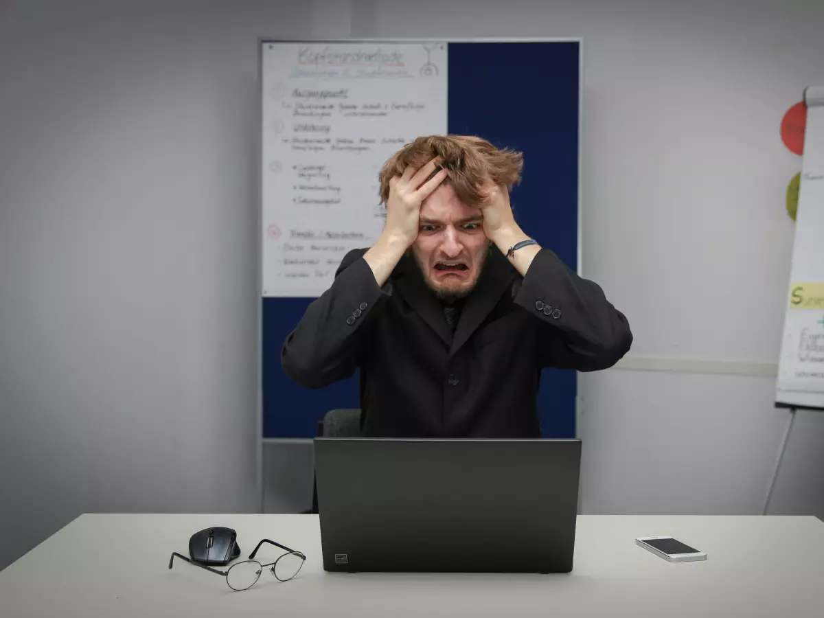 A young man sits in front of a laptop, clutching his head in frustration. He appears to be struggling with something on the computer. The background is a typical office setting.