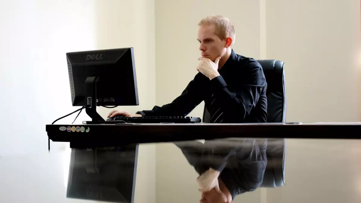 A man sits at a desk in front of a computer, staring at the screen. His reflection is visible on the glossy desk.