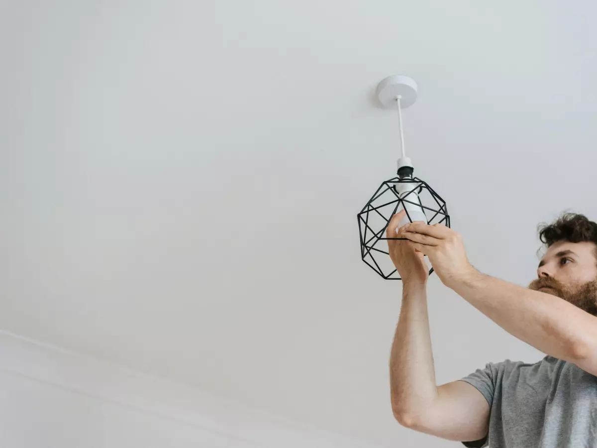 A man is seen from below, standing on a stepladder and replacing a light bulb on the ceiling. The lightbulb is a modern design with a metal cage.