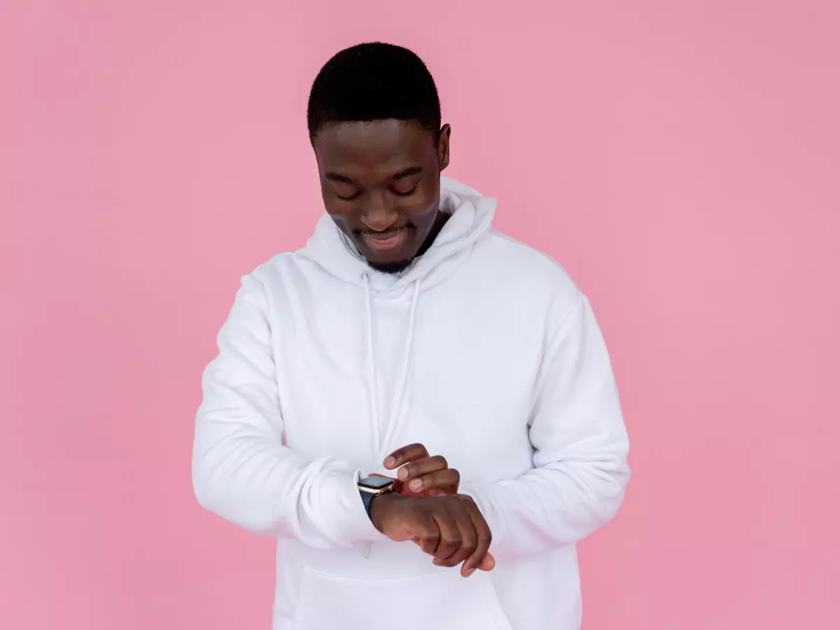 A young black man wearing a white hoodie looks down at his wrist checking his smartwatch. He is standing against a pink background.