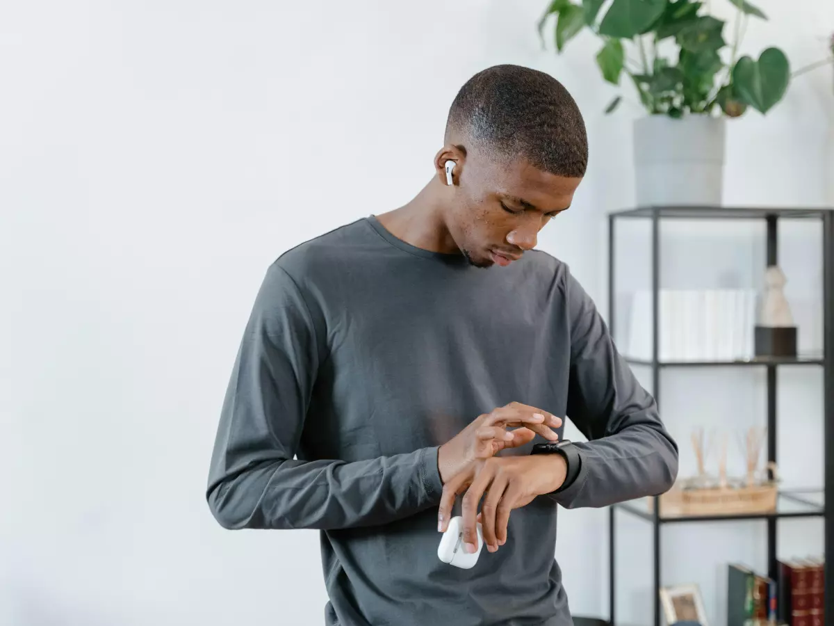 A man wearing a grey long-sleeved shirt is standing in a room and looking at his Apple Watch.