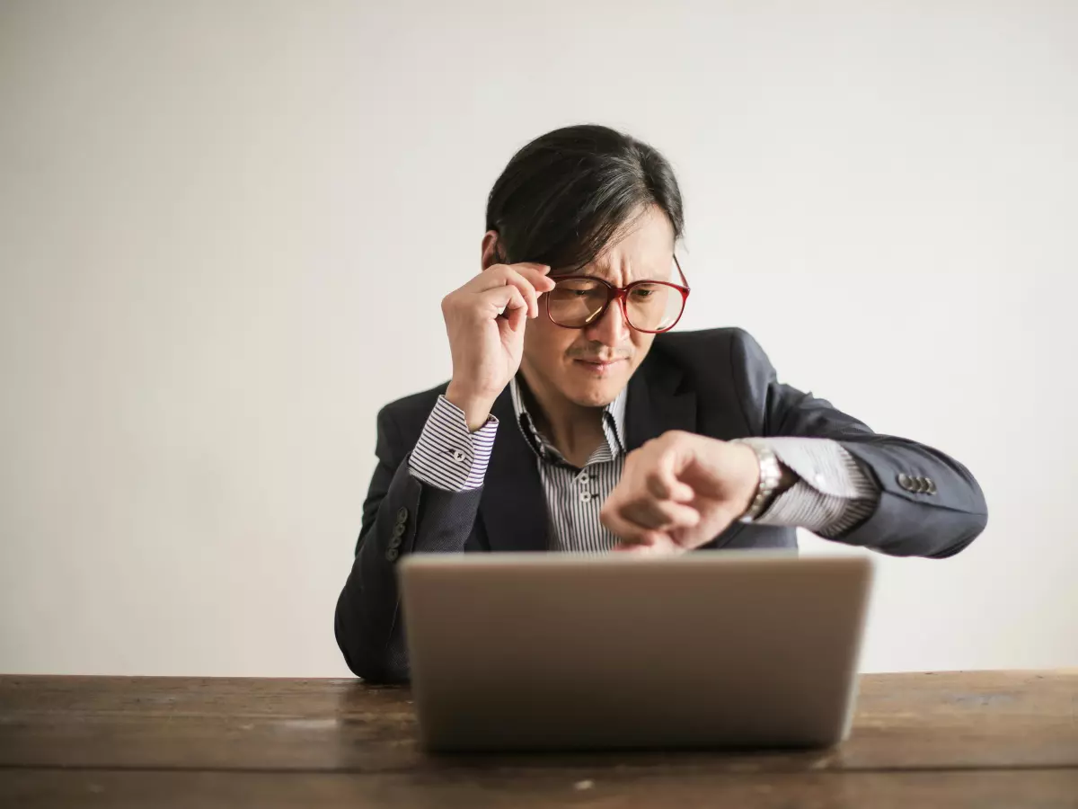 A man with dark hair is sitting at a desk in front of a laptop, wearing a suit and glasses. He is looking at the laptop with a worried expression.