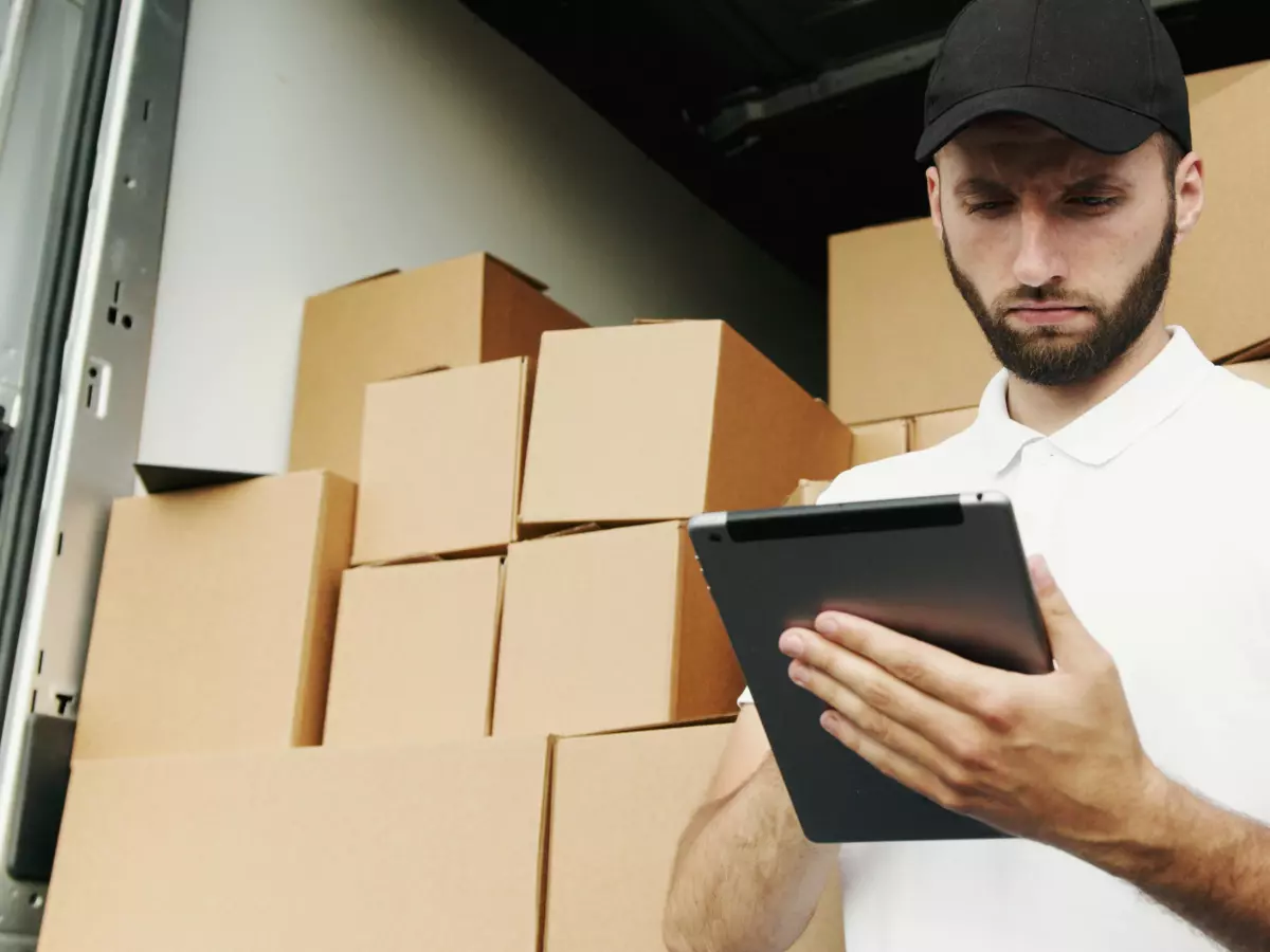 A man wearing a white shirt and a black cap is standing in the back of a delivery truck, surrounded by boxes. He is looking at a black tablet in his hands.