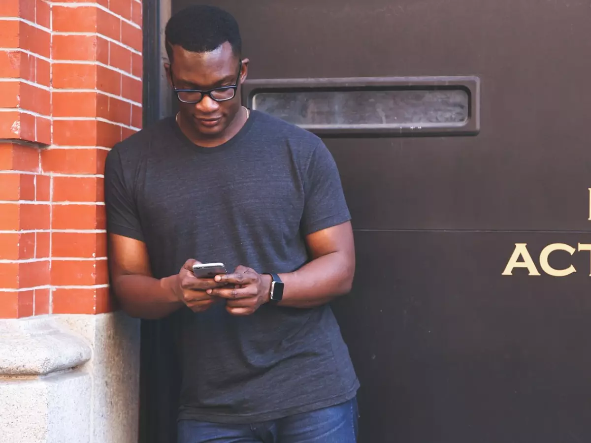 A man with glasses is standing outside a building and looking at his phone. The background is a brick wall and a black door with the words "NO ACTIVE" on it.