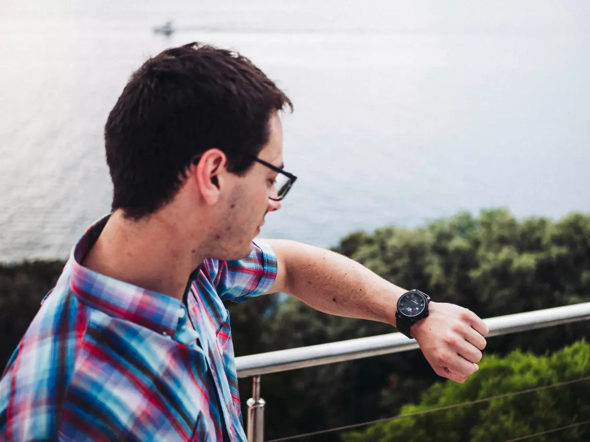 A man with glasses wearing a colorful shirt is looking at his watch while standing on a balcony overlooking the sea. The sky is blue and there are some green trees in the background. The man has a worried expression on his face.