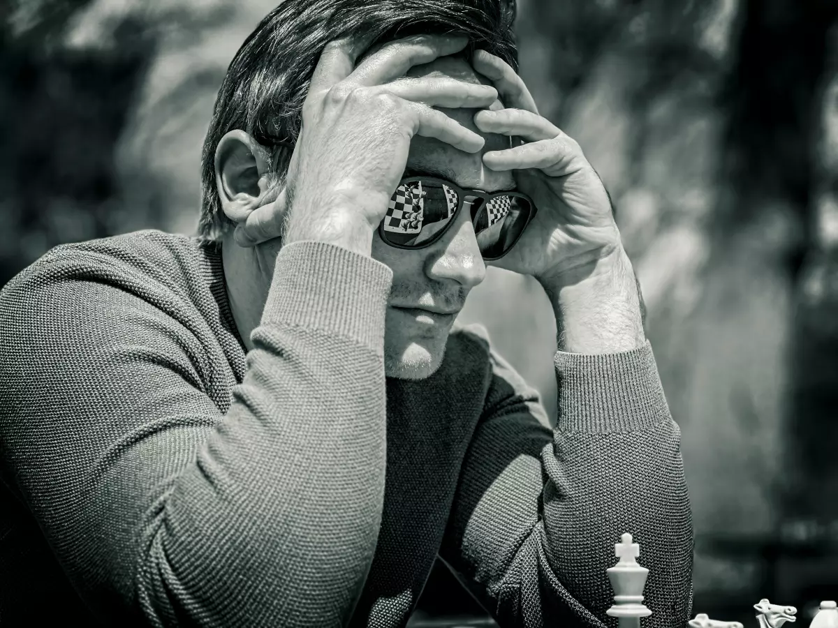 A man with his head in his hands, wearing sunglasses that reflect a chessboard, sits at a chessboard outdoors.