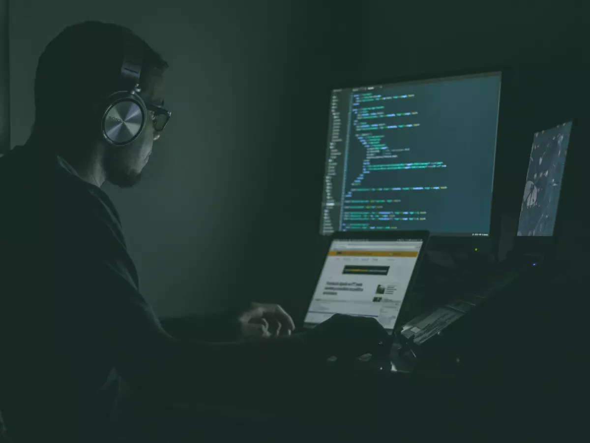 A person in a dark room, wearing headphones, works on a computer with code displayed on the screen.