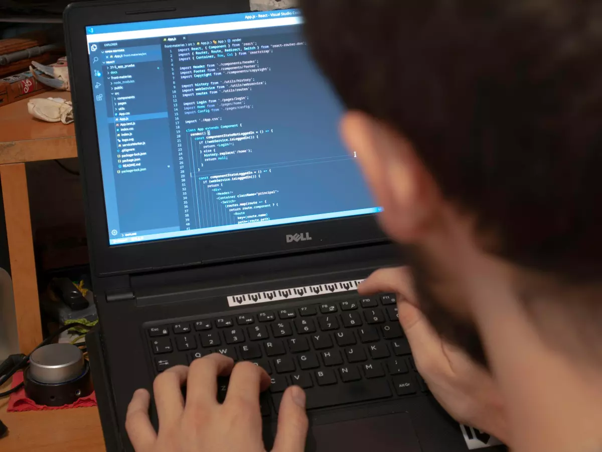 A man is typing on a laptop. The laptop has a blue screen with code on it. The man's hands are visible, and the laptop is on a desk. The background is blurred.