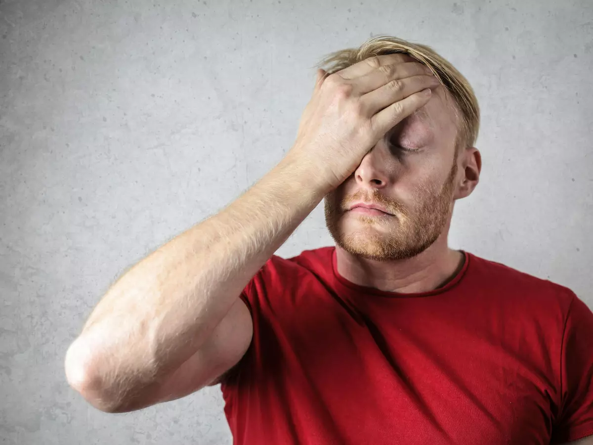 A man with blond hair and a beard is covering his face with his hand. He is wearing a red t-shirt. He looks distraught.